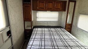 A compact RV bedroom with a bed, wooden cabinets, a window with blinds, and a ceiling vent.