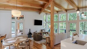 A spacious living area with a fireplace, wooden beams, large windows, and a modern kitchen. - The Briar House-A modern retreat in Hocking Hills (McArthur)