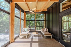 A wooden deck with two sofas and a table, large windows offering a view of trees, and a ceiling fan.