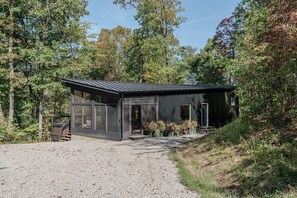 Eine moderne, dunkel gefärbte Holzhütte mit großen Fenstern, einer Schotterauffahrt und von dichtem Wald umgeben.