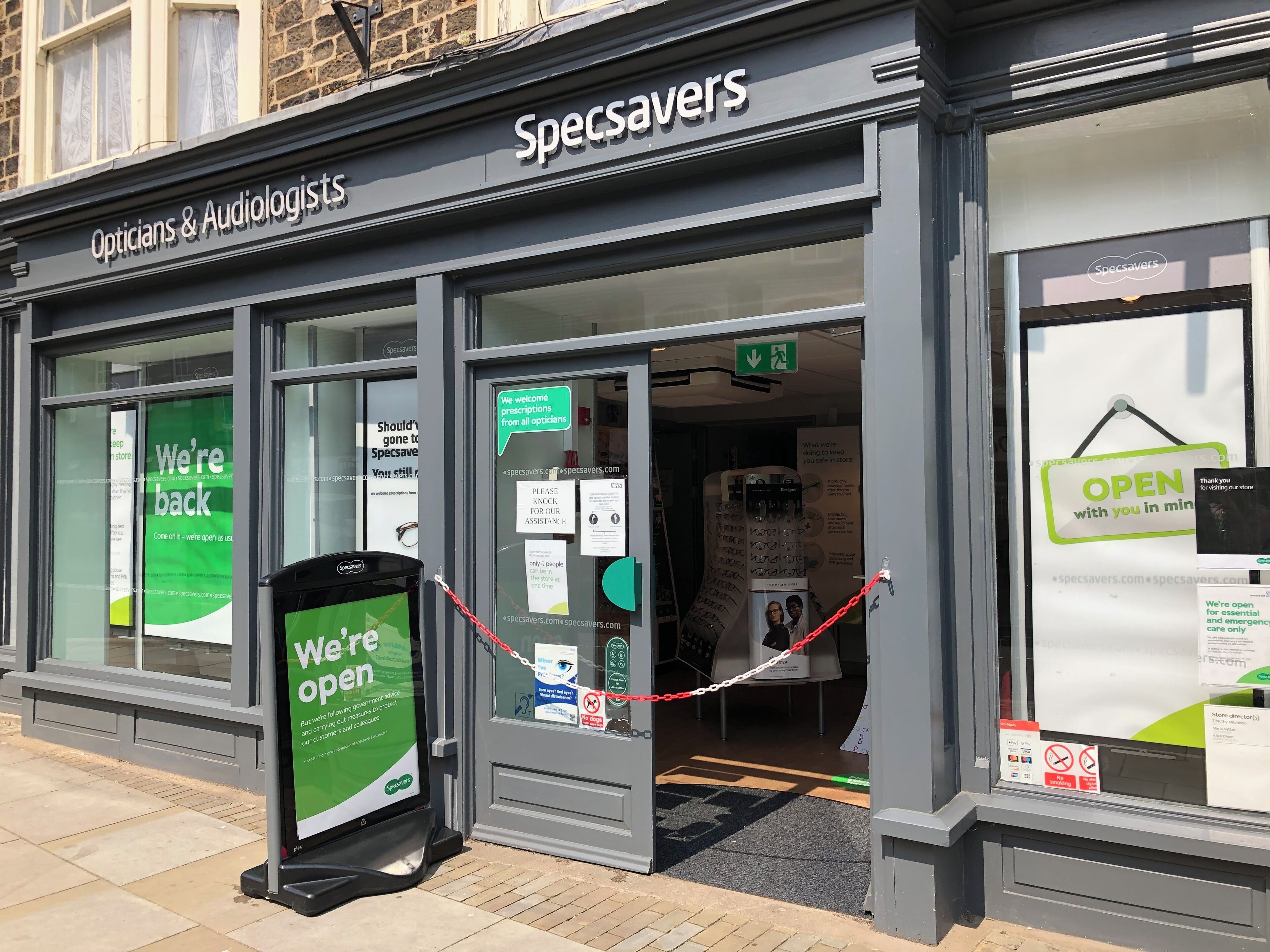 A Specsavers store with a green 'We're open' sign, a closed entrance, and a 'We welcome prescriptions from all opticians' notice.