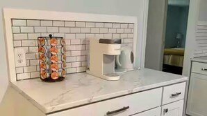 Une cuisine avec des meubles blancs, une machine à café et un présentoir à fruits.