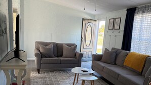 Un salon avec un canapé gris, une table basse blanche et un divan gris agrémenté d’un plaid jaune. On y trouve également un ventilateur de plafond, un téléviseur et un tapis à motifs.