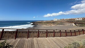 A coastal view with a wooden boardwalk leading to a beach, a rocky cliff, and buildings in the background. - La Esterlicia (Los Abrigos)