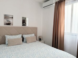 Une chambre avec un grand lit, deux tableaux accrochés au mur, un climatiseur, une table de chevet et une fenêtre avec des rideaux.