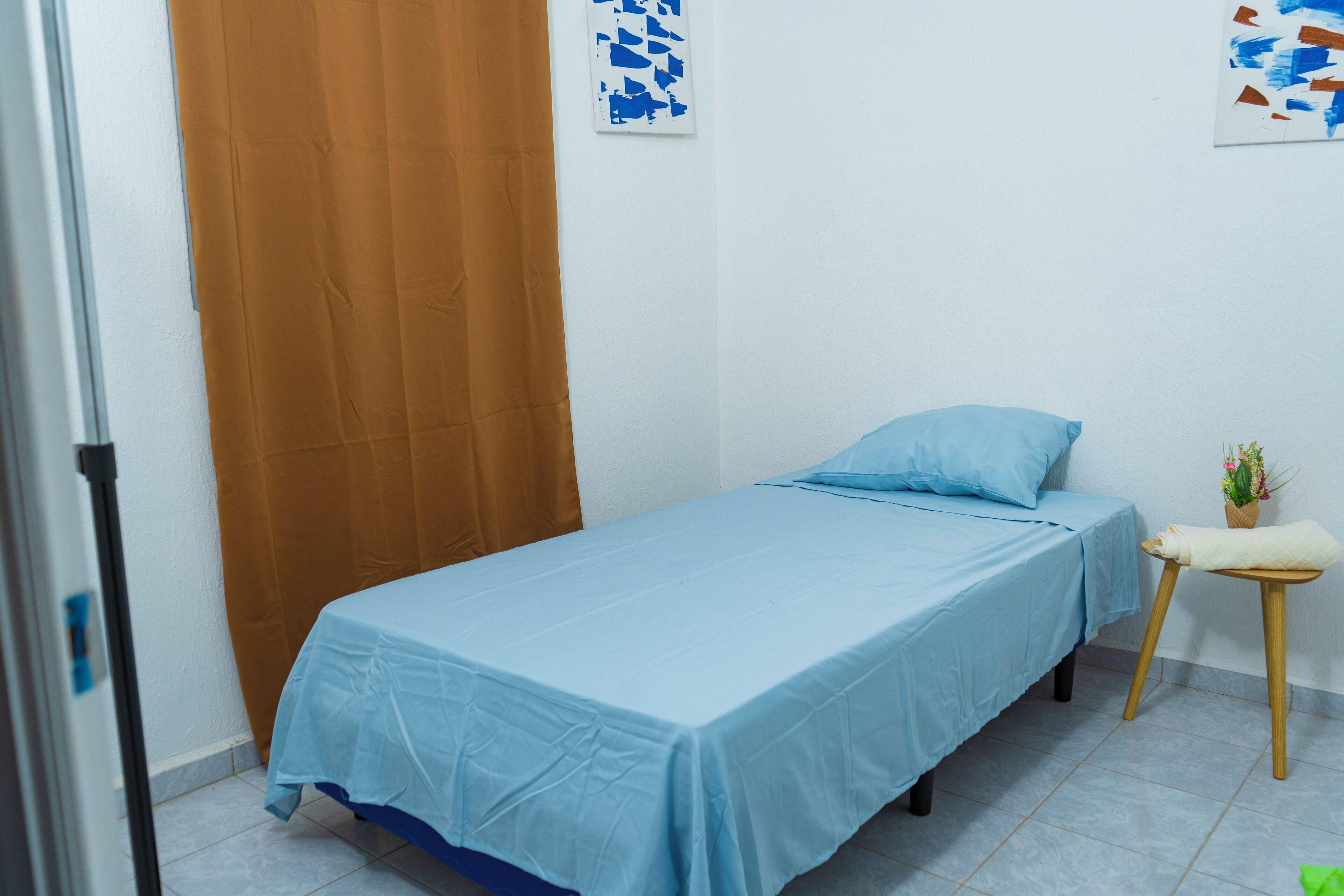 A single bed with a blue bedspread, a wooden side table with a plant, and a brown curtain.