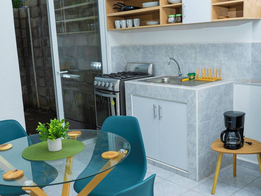 A modern kitchen with a dining area, featuring a glass table, teal chairs, and a small coffee table.