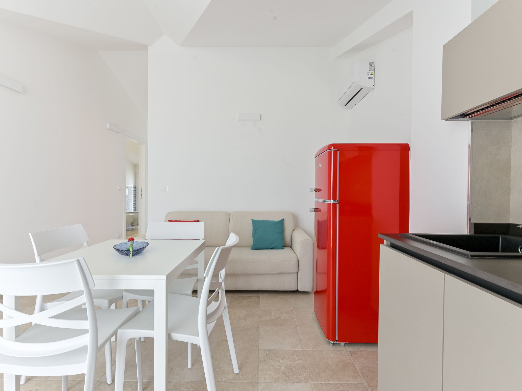 Une cuisine moderne avec des meubles blancs, un réfrigérateur rouge et un coin repas comprenant des chaises et une table, le tout dans des tons blancs.