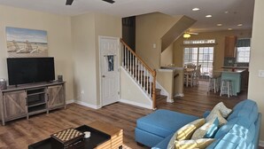 A spacious living room with a blue sofa, wooden entertainment unit, and a flat-screen TV. A staircase leads to a dining area with a kitchen in the background. - 4bedroom Beach Retreat in charming Long Neck with Rooftop View and lounging deck (Long Neck)