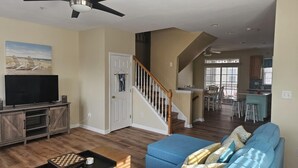 A spacious living room with a flat-screen TV, a wooden entertainment center, and a blue sofa. A staircase leads to a kitchen area with a bar and dining space. - 4bedroom Beach Retreat in charming Long Neck with Rooftop View and lounging deck (Long Neck)