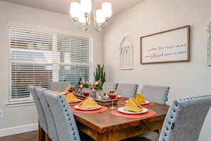 Une salle à manger avec une table en bois dressée pour six personnes, des chaises à revêtement gris et un lustre.