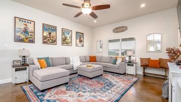 A living room with a sectional sofa, ottoman, patterned rug, ceiling fan, wall-mounted artwork, and a window with blinds.