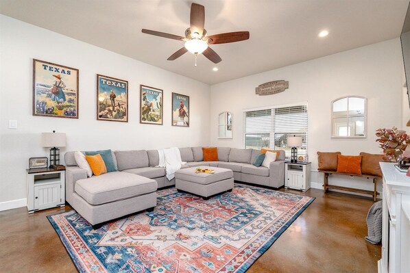 A living room with a sectional sofa, ottoman, patterned rug, ceiling fan, wall-mounted artwork, and a window with blinds.