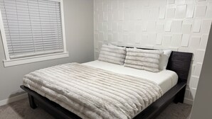 A bed with a white quilt and pillows, a dark wooden frame, and a window with blinds. - Fun, Cosy, Theater Basement! (Lehi)