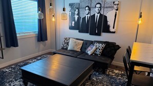 A modern living room with a dark sofa, a coffee table, and a dining table. There are wall art pieces and pendant lights. - Fun, Cosy, Theater Basement! (Lehi)