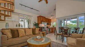 A spacious living area with a sofa, coffee table, and dining chairs. A kitchen with wooden cabinets and a dining area with a glass table and chairs are visible.