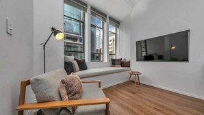 Ein modernes Wohnzimmer mit einer Couch, einem kleinen Tisch und einer Stehlampe. Große Fenster bieten einen Blick auf die Stadtlandschaft.