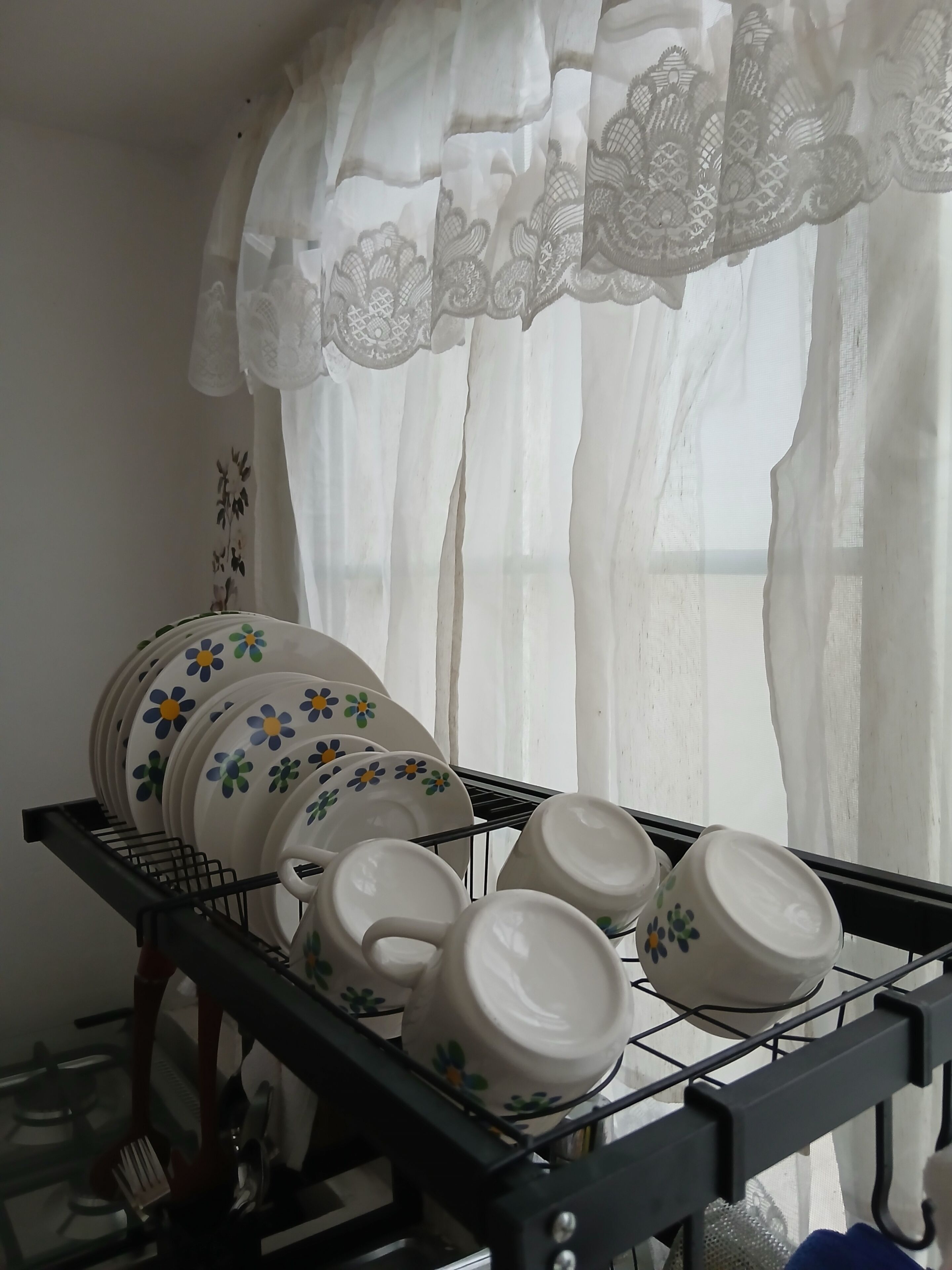 A kitchen area with a dish rack holding plates and cups, a gas stove, and a window with sheer curtains.