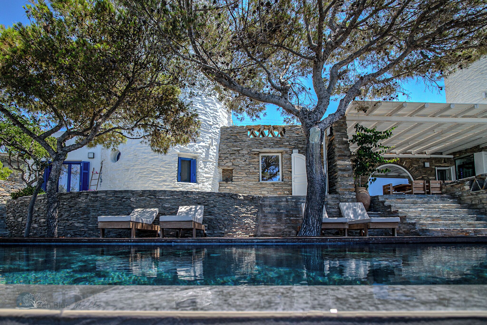 A poolside area with lounge chairs, a stone wall, and a building with a stone facade and blue shutters.