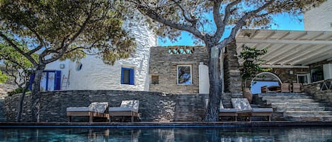 Zona junto a la piscina con sillones de descanso, pared de piedra y un edificio con fachada de piedra y contraventanas azules.