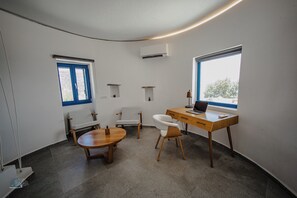 A modern room with a curved ceiling, a wooden desk with a laptop, a white chair, a small round table, and a window with a view of trees. - Zen Blue Suites Luxury and Nature in Kea (Kea)