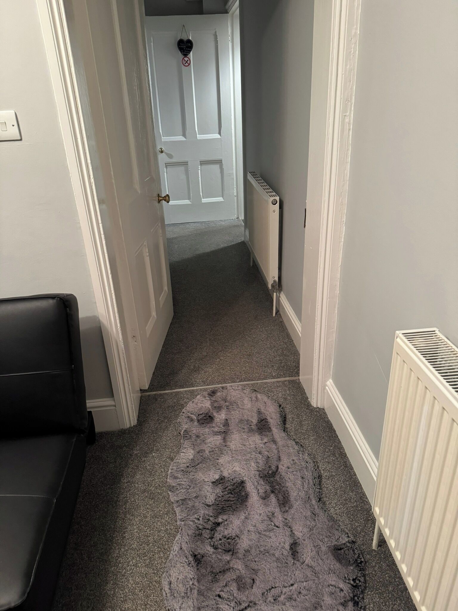 A narrow hallway with a grey carpet, a radiator, and a white door at the end.