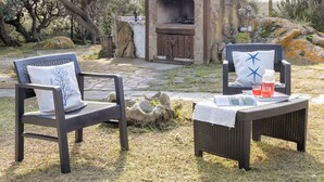 Deux fauteuils en osier avec des coussins blancs, une petite table recouverte d’une nappe blanche et un four extérieur en pierre.