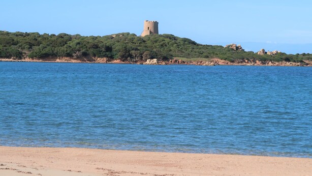 Une vue sur la côte, avec une tour perchée sur une colline, une eau d’un bleu limpide et une plage de sable.