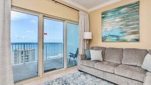A room with a sofa, a chair, a painting, and a view of the sea through a sliding glass door.