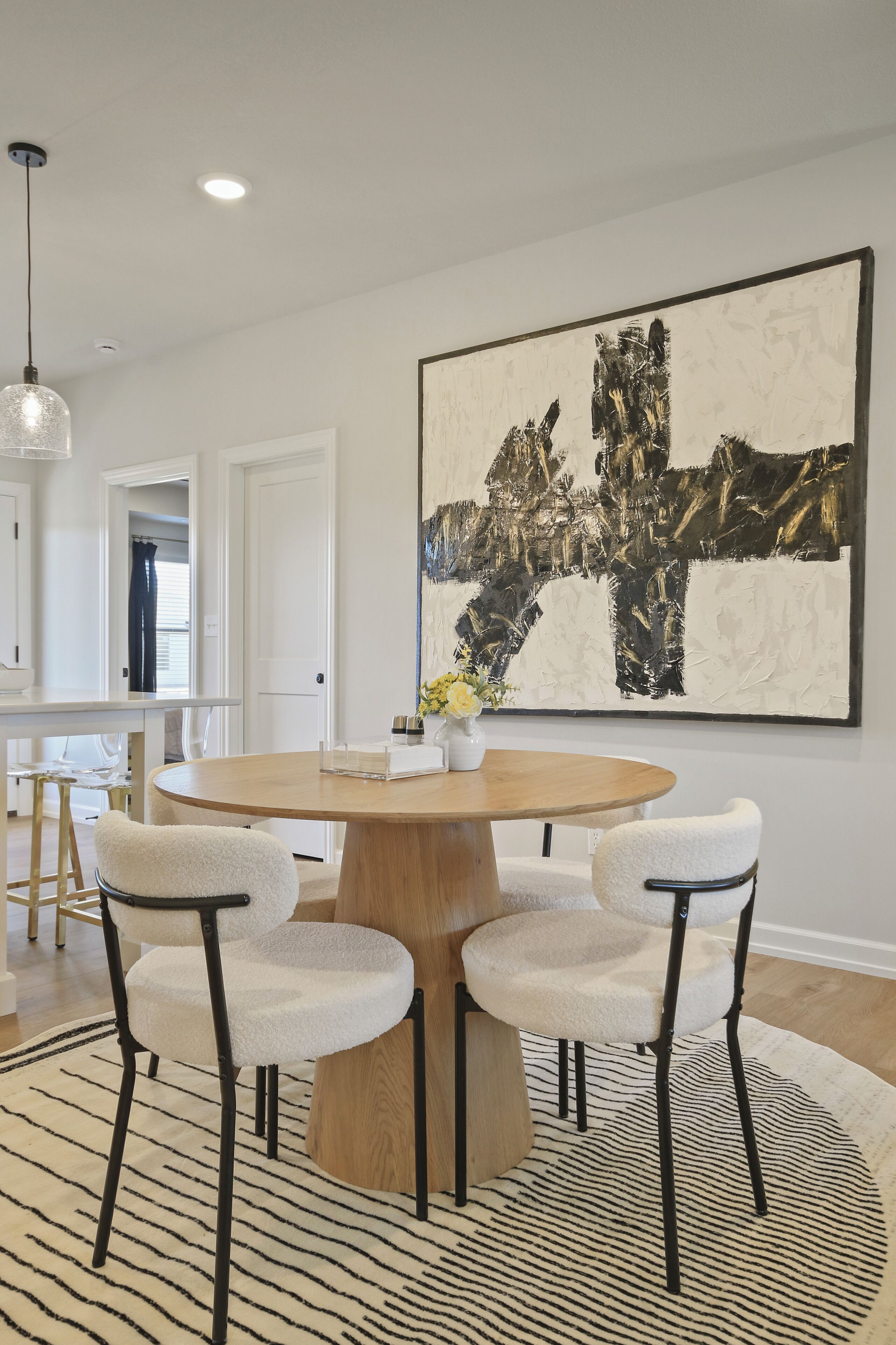 A dining area with a round wooden table, four white chairs, a large abstract painting, and a hanging light fixture.
