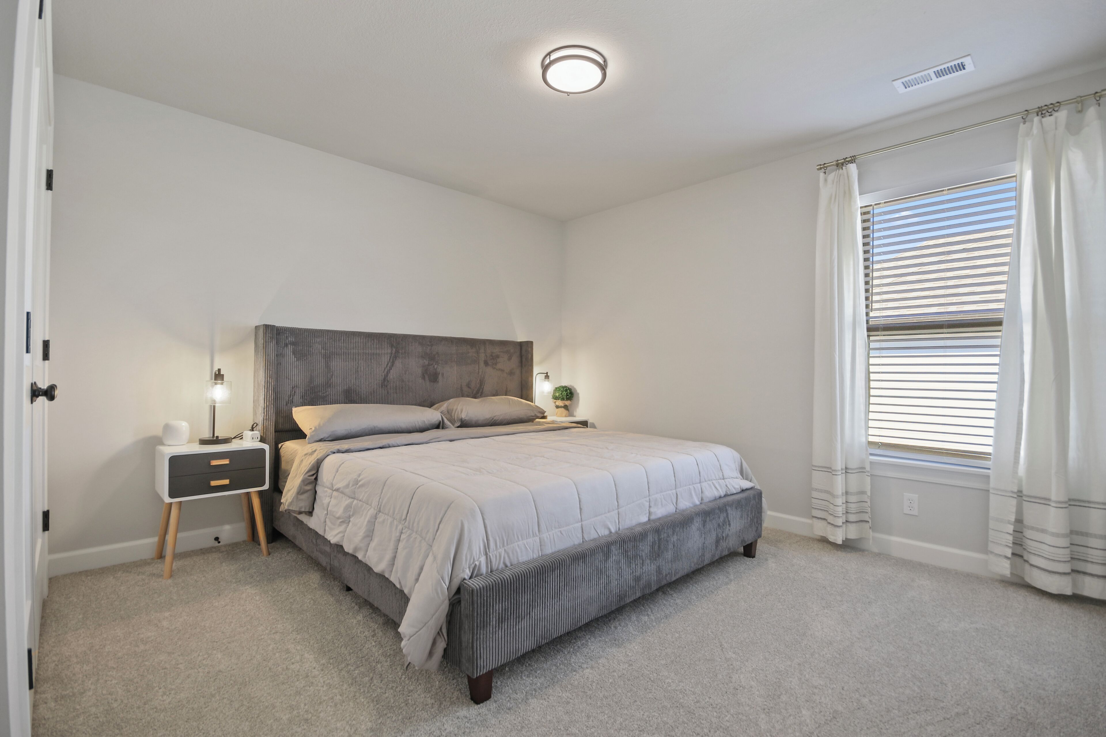 Une chambre à coucher avec un lit, des tables de chevet, une commode, une lampe et une fenêtre avec des rideaux.