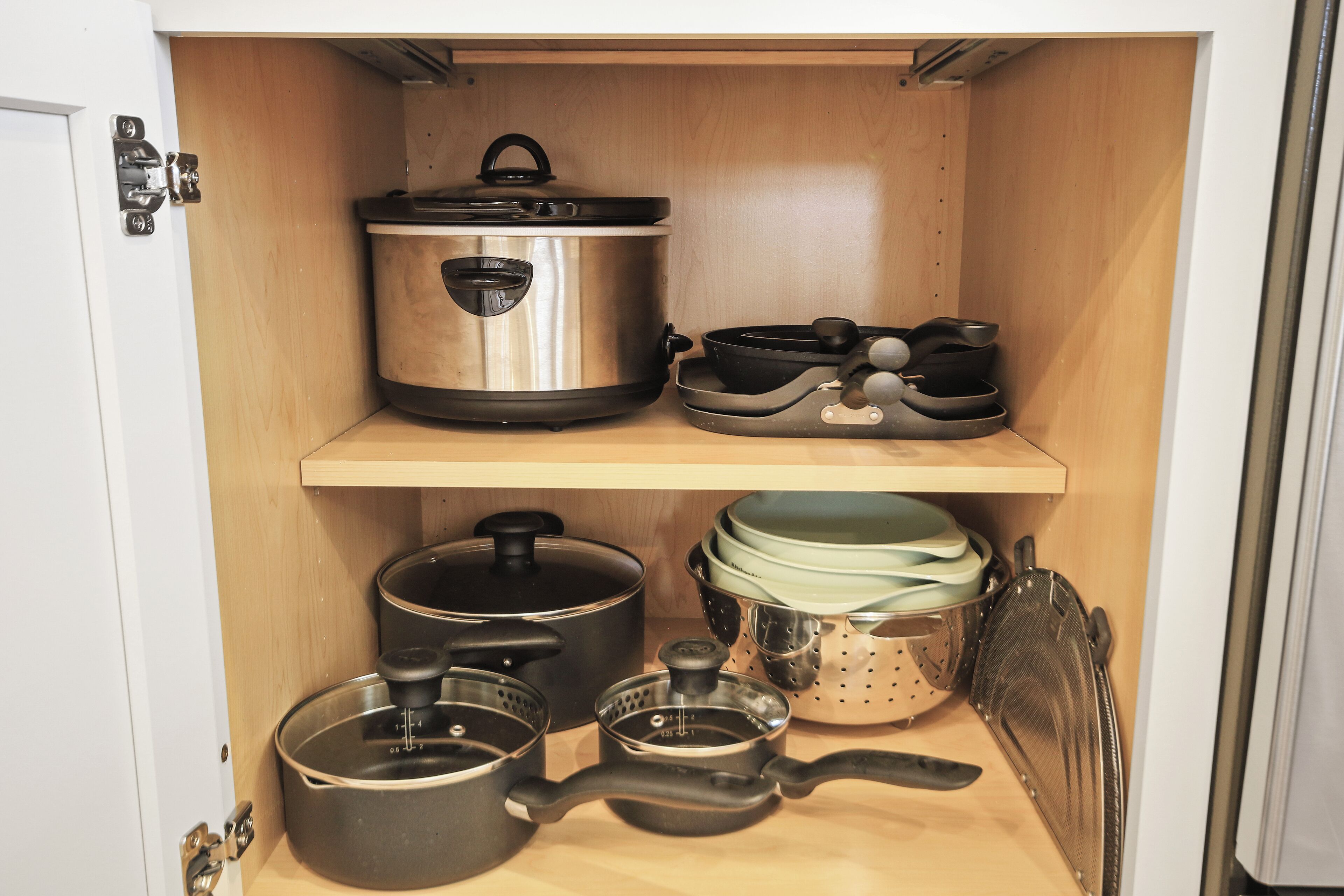 A kitchen cabinet with stainless steel cookware, including pots, pans, and a colander.