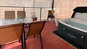 Una habitación con una mesa de vidrio, sillas de mimbre, un sofá negro y una mesita con botellas y una planta.