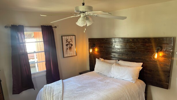Un dormitorio con una cama grande, un ventilador de techo, una ventana con cortinas y una lámpara de pared.