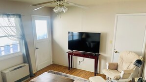 Una sala de estar con una televisión sobre un mueble de madera, un ventilador de techo, un sillón blanco, una mesita y un tapete estampado.