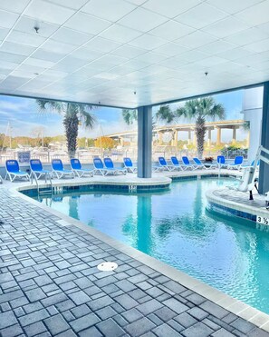 Una piscina interior con agua cristalina, rodeada de sillas reclinables azules y palmeras.