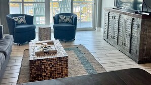 A modern living room with a large TV, two armchairs, a coffee table, and a view of a cityscape through large windows.