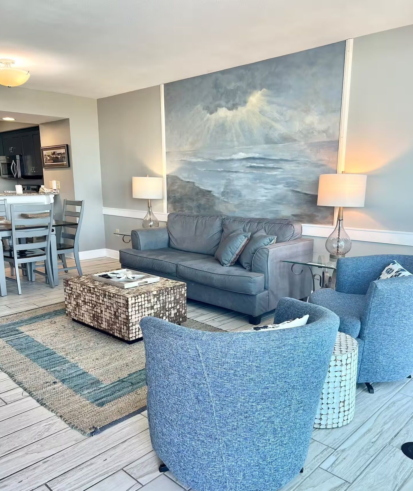 Un salon moderne avec un canapé gris, des fauteuils bleus et une table basse. Un tableau représentant un paysage marin est accroché au mur, et on aperçoit un coin repas à l’arrière-plan.