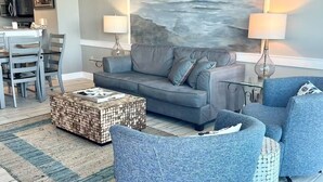 A modern living room with a grey sofa, blue armchairs, and a coffee table. There is a painting of a seascape on the wall, and a dining area is visible in the background.