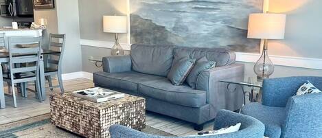 Un salon moderne avec un canapé gris, des fauteuils bleus et une table basse. Un tableau représentant un paysage marin est accroché au mur, et on aperçoit un coin repas à l’arrière-plan.