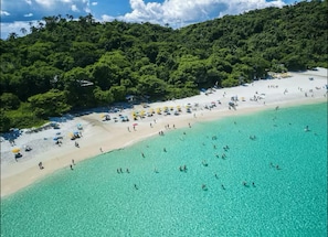 Vista aérea de una playa con aguas turquesas cristalinas, una orilla arenosa y colinas verdes exuberantes al fondo.