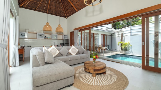 A modern living room with a grey sectional sofa, a wooden coffee table, and a pool area visible through glass doors. - Elephant Villa 5 by Villa Finder (Ubud)