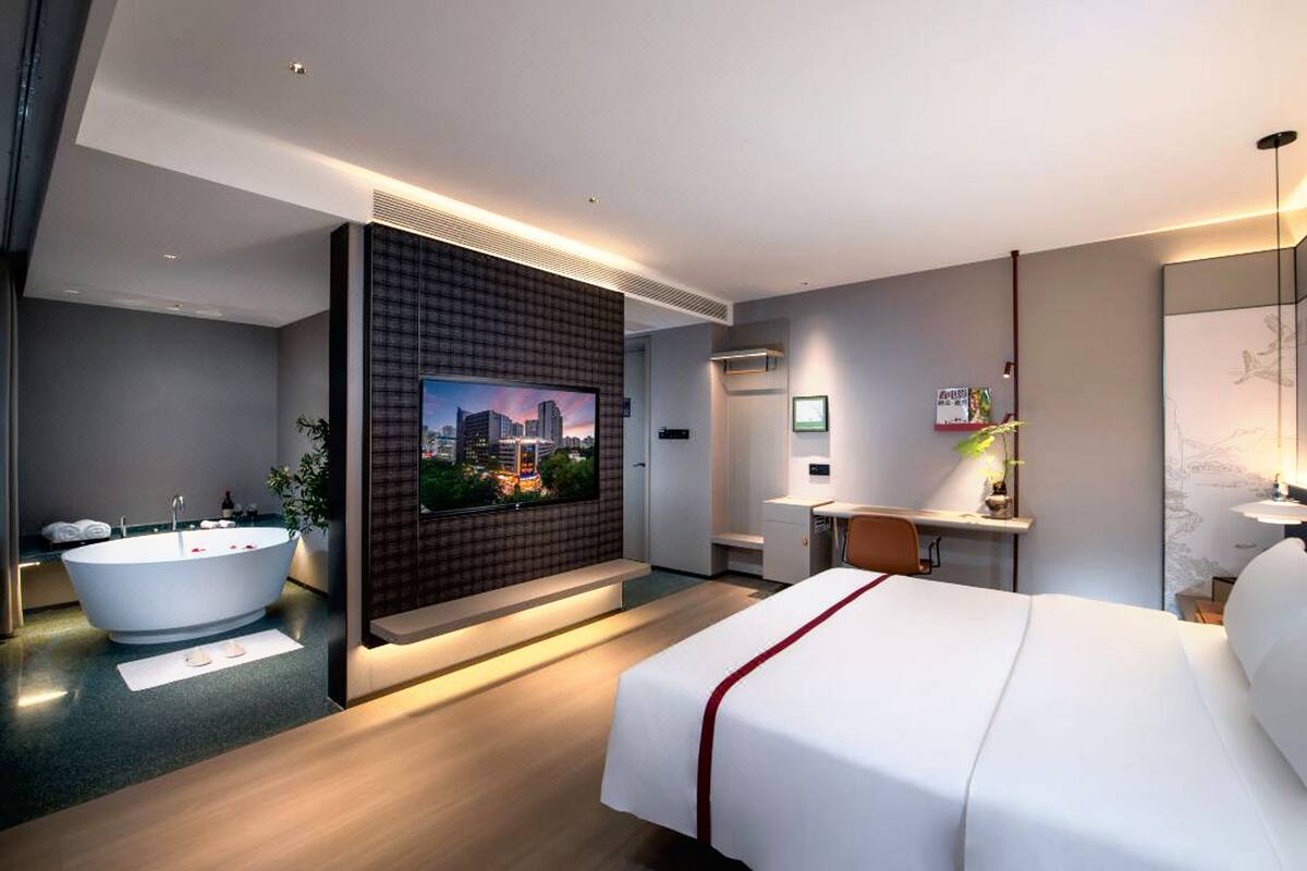 A modern hotel room with a large bed, a bathtub, a desk, and a TV displaying a cityscape.