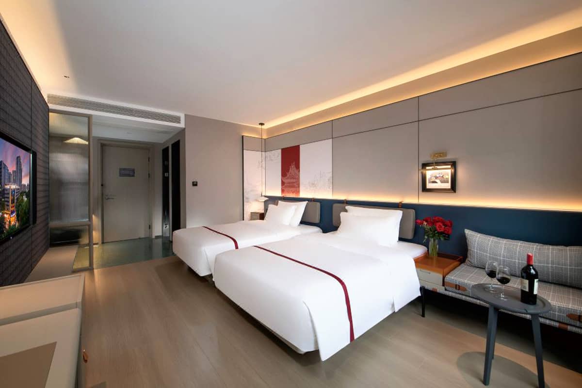 A modern hotel room with two beds, a small table with wine glasses and a bottle, and a sofa.
