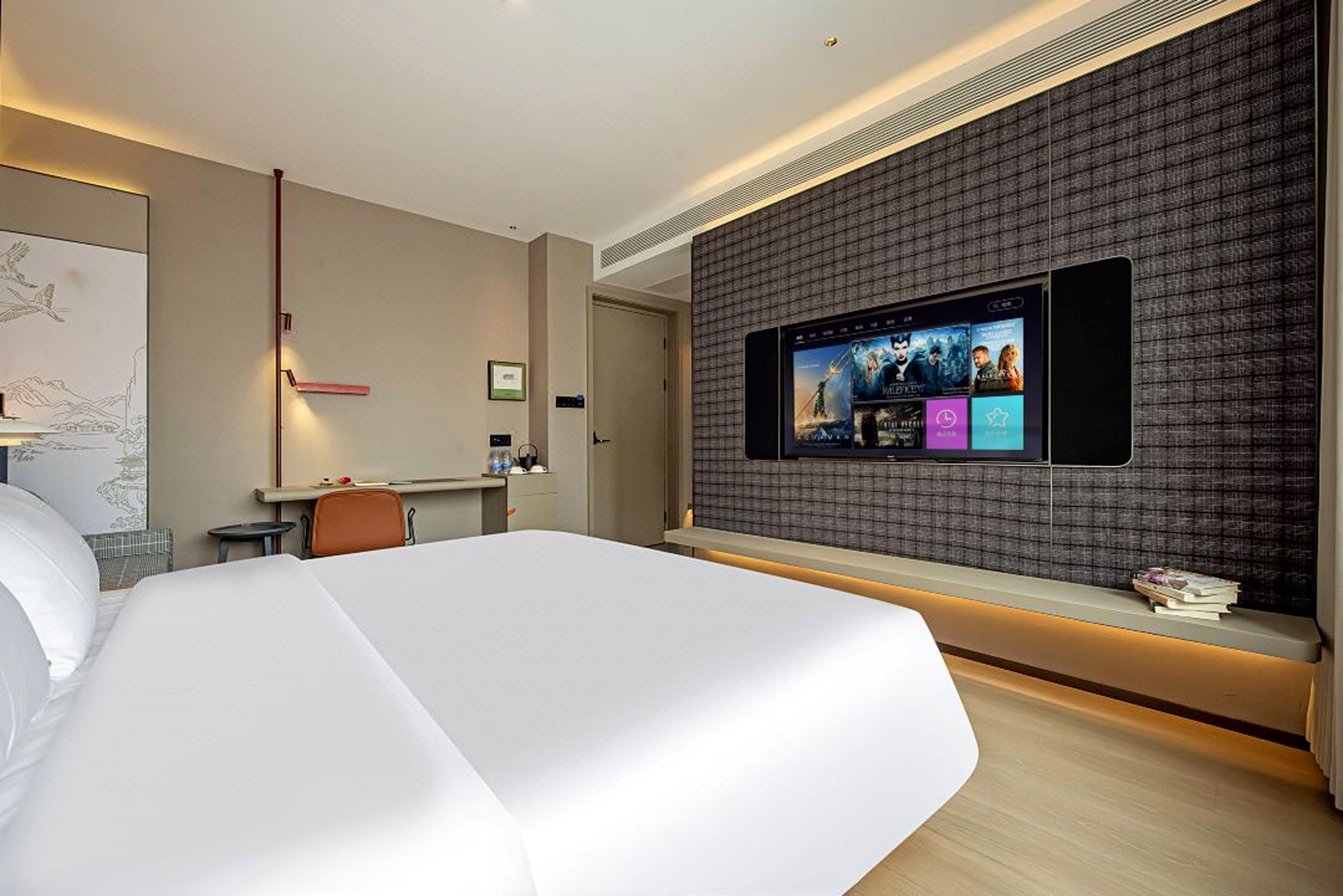 A modern hotel room with a large bed, a flat-screen TV mounted on the wall, a desk, and a seating area with a bench.