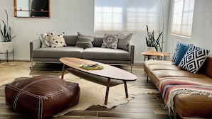 A modern living room with a grey sofa, a brown leather ottoman, a wooden coffee table, and a beige rug. There are two side tables with lamps, a large window with blinds, and a flat-screen TV mounted on the wall. - Charming 3-bedroom house with Pool/Jacuzzi and Desert Mountain View (Desert Hot Springs)