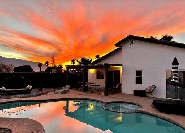 A swimming pool with a sunset view, a covered patio area, and a white building with a flat roof. - Charming 3-bedroom house with Pool/Jacuzzi and Desert Mountain View (Desert Hot Springs)