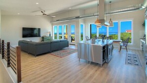 Une cuisine moderne avec un îlot central, de grandes fenêtres offrant une vue sur l’océan, un canapé gris et un ventilateur de plafond.