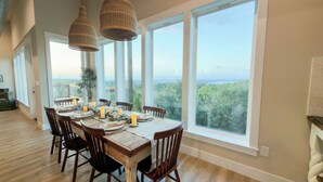 Un espace repas avec une grande table dressée pour six personnes, de grandes fenêtres offrant une vue sur le paysage et des lampes en osier suspendues.
