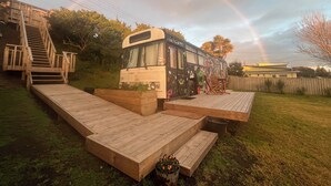 A bus with a rainbow in the background, wooden steps leading up to a deck, and a chair on the deck. - Luxury Beach Bus (Whanganui)